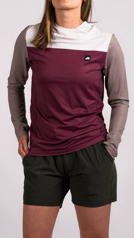A person in a long-sleeve Sun Hoodie by Belong Designs, featuring a color-blocked design with white, maroon, and gray sections, a black logo on the chest, and wearing dark green shorts stands with hands in pockets. The background is plain white.